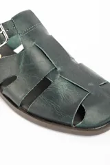 Zueco tipo cangrejera de cuero lavado color verde oscuro, con diseño de tiras entrelazadas y hebilla metálica lateral ajustable.