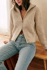 Cardigan de punto acanalado en color beige melange con motas oscuras. Presenta un diseño ajustado al cuerpo, cierre frontal completo con cremallera metálica y cuello alto.