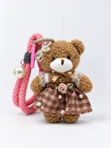 Llavero en forma de osito de peluche con vestido a cuadros rosado y marrón, adornado con encaje, perlas y una rosa de tela. Incluye lentes dorados, moño en el cuello, detalle de perlas en la cabeza y una correa de mano trenzada de color rosado con una cuenta perlada colgante.