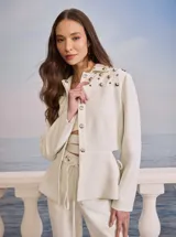 Chaqueta corta color blanco, con cuello camisero, cierre frontal con botones metálicos y apliques de tachas metálicas en los hombros.