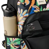 Mochila Rip Curl Ozone 2.0 de 30 litros, con estampado tropical multicolor sobre fondo negro. Cuenta con compartimento principal con cierre, bolsillo frontal organizador, correas ajustables y acolchadas, y compartimento interno para laptop.