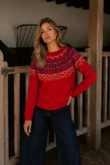 Buzo de lana merino con diseño jacquard en el canesú, presentando motivos geométricos en tonos azul, blanco y mostaza sobre una base roja. Posee cuello redondo, mangas largas y puños y ruedo acanalados.