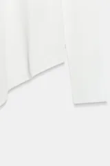Jersey de punto liso blanco, con cuello redondo y mangas largas. Presenta un diseño de bajo asimétrico pronunciado.