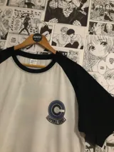Remera blanca con mangas negras y cuello redondo. Estampado frontal con el logo de Capsule Corp de la serie Dragon Ball.