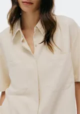 Camisa de manga corta color beige, con cuello camisero, cierre frontal de botones y dos bolsillos delanteros con solapa.