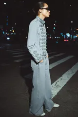 Chaqueta de denim con efecto lavado, corte entallado y cuello redondo. Presenta un diseño de estilo militar con cierre frontal de cremallera metálica y aplicaciones decorativas de botones y costuras en el delantero y los puños.