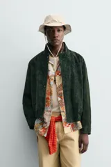 Cazadora regular fit de piel con acabado en ante color verde oscuro, de la colección especial Gabriel Azevedo x ZARA. Cuenta con cuello subido, manga larga, bolsillos de vivo en la cadera, bolsillo interior y bajo acabado con elástico. Cierre frontal con cremallera oculta por solapa con botones a presión.