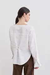 Blusa de lino con cuello bote y mangas largas terminadas en puños acampanados con volados. Presenta un cierre con botón en la espalda.