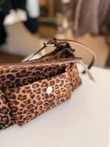Cartera tipo baguette con estampado animal print, confeccionada en material suave símil gamuza. Presenta un bolsillo frontal con solapa y cierre, compartimento principal con cierre superior, y asa larga regulable de cuero sintético marrón.