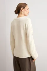 Sweater de punto color blanco natural, con cuello redondo y mangas largas adornadas con botones dorados a lo largo de las costuras exteriores.