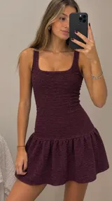 Vestido corto color bordó, con escote cuadrado y tirantes anchos. Presenta un diseño ajustado al cuerpo con costuras verticales y una falda con volado en el ruedo, confeccionado en tela con textura arrugada.
