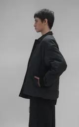 Campera tipo sobrecamisa de diseño oversize, confeccionada en microfibra opaca con patrón matelaseado geométrico. Presenta cuello, puños, cartera central y ruedo en gabardina de algodón, con cierre frontal mediante broches a presión. El diseño del ruedo simula una prenda superpuesta.