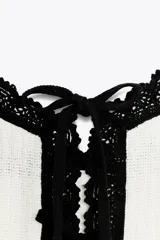 Top corto de tejido acanalado color blanco, con escote pico, tirantes finos negros y ribetes de encaje negro en el escote y el ruedo. Presenta un cierre frontal con lazada ajustable.