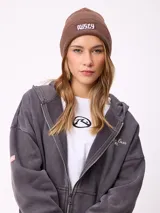 Gorro tipo beanie de tejido de punto acanalado en color marrón, con parche rectangular frontal con el logo de la marca Rusty.