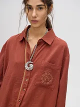 Camisa de corte holgado color azul petróleo, con cuello clásico, cierre frontal de botones y bolsillo cuadrado en el pecho con logo bordado. Presenta detalles de ribetes en contraste en la cartera de botones y en los puños.