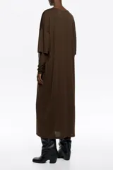 Vestido largo de corte holgado en color marrón, con cuello redondo y mangas largas de tul superpuestas. Presenta un diseño minimalista con sutiles detalles brillantes en el tejido principal.