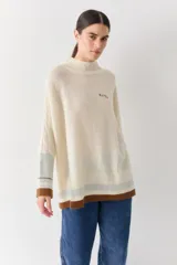 Buzo tejido de silueta amplia con cuello alto, mangas largas con detalle de trenzado y puños en contraste. Presenta un diseño con franjas horizontales en la parte inferior y el logo de la marca bordado en el pecho.