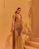 Vestido largo de tejido crochet en color bronce, con escote en V profundo, diseño de red y detalles trenzados. Presenta una silueta ajustada y un acabado con flecos largos en el ruedo.