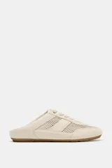 Zapato deportivo tipo mule, color crema o beige claro, con diseño destalonado. Presenta paneles de malla transpirable combinados con apliques de cuero liso y texturizado, y cordones en la parte delantera. La suela es plana de goma color caramelo.