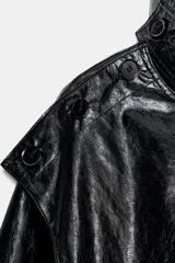 Chaqueta de efecto piel en color negro con cuello subido y bufanda desmontable integrada mediante botones. Presenta manga larga con trabillas ajustables en los puños, cierre frontal con cremallera y bolsillos laterales ocultos en las costuras. El diseño incluye un bajo con elástico en la espalda y forro interior.