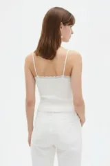 Musculosa de punto blanco, corte crop, con tirantes finos, escote en V adornado con detalles de encaje y un pequeño lazo en el centro.