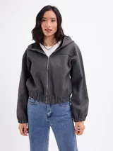 Campera de paño color beige con capucha, cierre frontal completo y mangas largas con puños elásticos. Presenta un diseño corto con ruedo elástico.