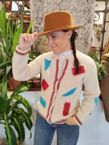 Cardigan de lana merino tejido a mano con técnica de dos agujas, de color marfil con parches geométricos de tela en tonos celestes y lila aplicados con costuras visibles. Presenta cierre frontal con botones de madera y detalles de ribetes multicolores en los bordes.