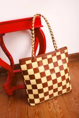 Bolso tipo tote de cuero trenzado a mano con diseño de damero en colores marrón y beige. Cuenta con doble asa trenzada y forro interior.