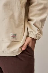 Campera canguro de gabardina color beige, con capucha ajustable con cordones, cierre frontal medio y bolsillo tipo ojal con solapa en el frente. Presenta mangas largas con puños elásticos y diseño holgado.