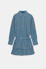 Vestido corto de manga larga con cuello solapa y cierre frontal de botones a presión. Presenta un diseño a cuadros en tonos azul y marrón, cintura ajustable con cinturón de hebilla y falda con volantes en el bajo.