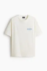 Remera de algodón de peso medio en color blanco, con cuello redondo, hombros caídos y corte holgado. Cuenta con un pequeño estampado ovalado azul con la palabra 'Uncover' y un símbolo en el pecho.