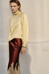 Blusa de satén color lila con cuello subido, manga larga con puños anchos y detalle de botones dorados en el hombro.