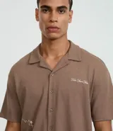Camisa de manga corta, color marrón topo, con cuello camisero y corte amplio (boxy). Presenta bordados minimalistas en blanco en el frente, simulando un paisaje.