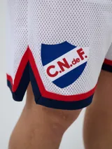 Short deportivo Umbro del Club Nacional de Football, modelo Match para hombre. Diseño en color azul marino con tejido de malla, detalles en blanco y rojo en los laterales y el escudo del club bordado.