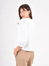 Camisa blanca de corte clásico con cuello redondo y cierre frontal oculto. Presenta mangas tres cuartos abullonadas con puños elásticos, brindando un toque femenino y moderno.