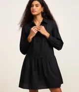 Vestido corto de tricolina color negro, con cuello camisero, cierre frontal de botones y mangas largas. Presenta un panel frontal con detalles bordados tipo broderie y falda con corte evasé.