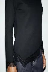 Top de manga larga en tejido suave color gris, con cuello redondo y bajo asimétrico decorado con encaje negro a tono. Presenta detalles de encaje negro en los puños.