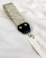 Llavero con cinta de tela estampada con diseño de cebra en tonos grises. Tiene un conector de cuero sintético negro con remaches metálicos y un gancho mosquetón plateado.