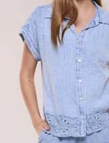 Blusa de algodón celeste con textura tramada tipo denim lavado, cuello solapa abierto en V, botones frontales y detalle de puntilla tejida al crochet en el ruedo.