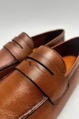 Mocasín de cuero color café, diseño clásico de estilo penny loafer con suela plana y costuras visibles.