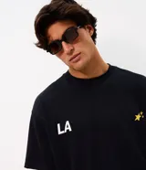 Remera de corte oversized confeccionada en mezcla de algodón y poliéster. Presenta cuello redondo, mangas cortas con hombros caídos y diseño estampado en la espalda con la frase 'LA THE START OF A NEW ERA' acompañada de estrellas amarillas.