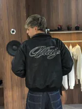 Campera de cuero sintético negro con diseño tipo bomber, cuello camisero y puños y ruedo elásticos. Presenta un bordado grande en la espalda con tipografía cursiva en color blanco.
