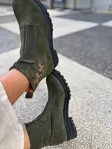 Botas cortas de cuero gamuzado color marrón oscuro, con suela track de goma negra y cierre frontal. Presentan un detalle de dos cruces bordadas a mano en tonos naranja y rojo en el lateral exterior.