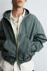 Cazadora negra de algodón con efecto lavado, corte relajado y capucha ajustable. Presenta cierre frontal de cremallera, bolsillos frontales tipo canguro y elásticos en puños y bajo.