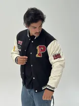 Campera estilo varsity con cuerpo negro y mangas blancas. Presenta parches bordados en el pecho y las mangas, cierre frontal con botones a presión y puños, cuello y ruedo acanalados con rayas blancas y negras.