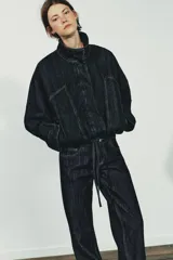 Cazadora de denim azul oscuro con cuello solapa, cierre frontal con cremallera y botones a presión ocultos. Presenta mangas largas tipo murciélago con puños, bolsillos de vivo en el delantero y bajo elástico ajustable con cordones.