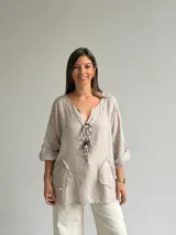 Blusa holgada de lino color beige, con escote en V, mangas tres cuartos con trabilla y bolsillos delanteros aplicados con bordes deshilachados. Cuenta con tiras frontales decorativas con botones de madera.