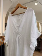 Vestido midi de lino color beige, con diseño oversize, escote en V y detalle de nudo drapeado en el frente. Posee mangas cortas amplias con dobladillo y corte asimétrico, siendo más largo en la parte trasera.