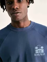 Camiseta azul de manga corta con cuello redondo y estampado frontal con palmeras y texto.