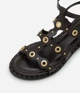 Sandalia plana tipo gladiadora color negro, confeccionada en poliuretano. Presenta tiras onduladas con recortes ahuecados y detalles de ojalillos y tachas metálicas doradas. Cuenta con doble tira ajustable en el tobillo con hebillas metálicas.
